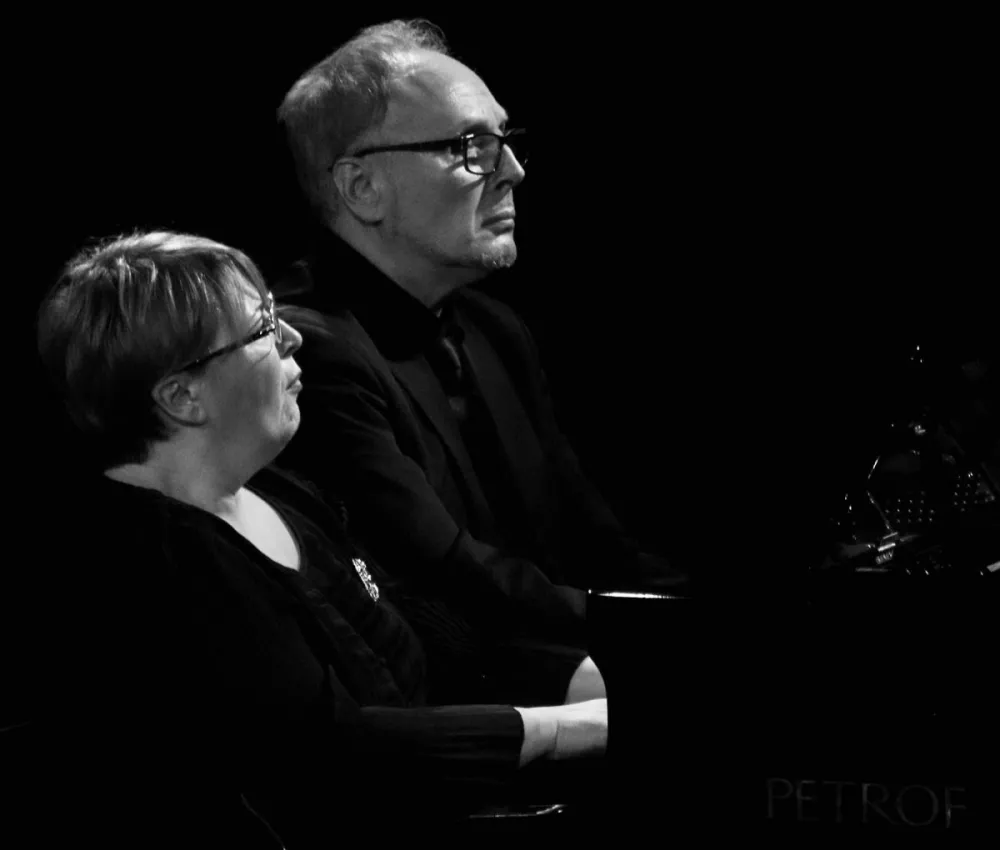 Czarno-biała fotografia dwojga pianistów grających razem na jednym fortepianie.